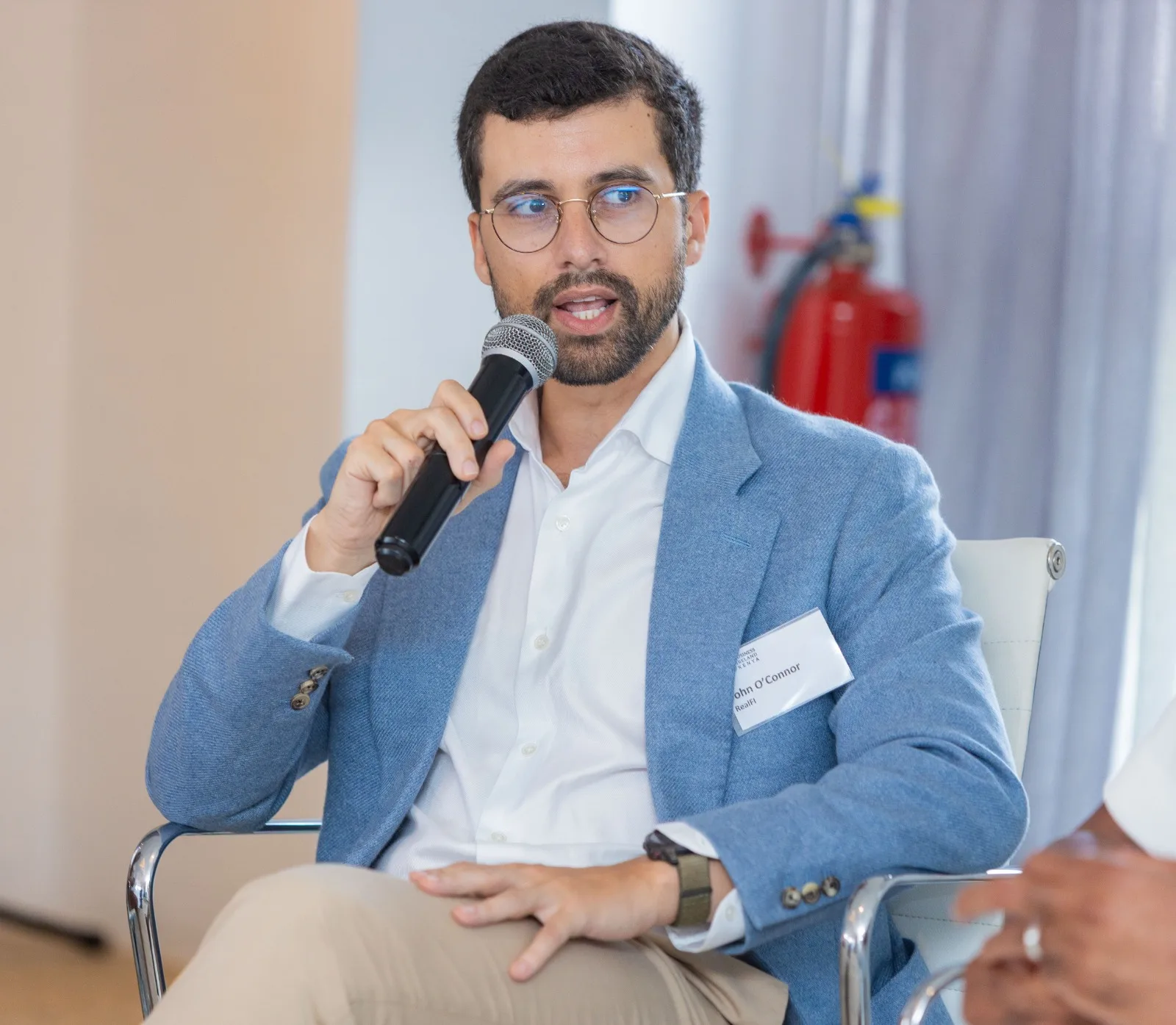 A seated speaker in a light blue jacket holding a microphone at a Chamber event